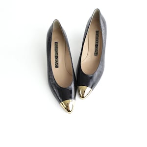 May include: Pair of black leather pumps with gold toe caps. The shoes have a pointed toe and a low heel. The brand name "Boutique Osaka" is printed on the inside of each shoe.