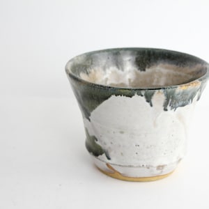 Può includere: Un piccolo vaso in ceramica bianca con una glassa verde e marrone. Il vaso ha un bordo svasato e una base arrotondata.