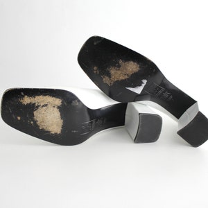 Puede incluir: Un par de mules blancas de tac&oacute;n con suelas negras. Los zapatos est&aacute;n colocados sobre una superficie blanca con las suelas hacia arriba.
