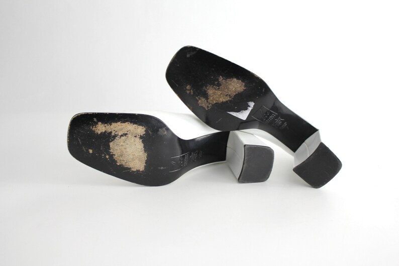 Puede incluir: Un par de mules blancas de tac&oacute;n con suelas negras. Los zapatos est&aacute;n colocados sobre una superficie blanca con las suelas hacia arriba.