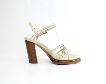 Puede incluir: Sandalias de tacón de cuero crema con plataforma de madera y múltiples tiras. Las sandalias tienen cierre de hebilla y punta puntiaguda.