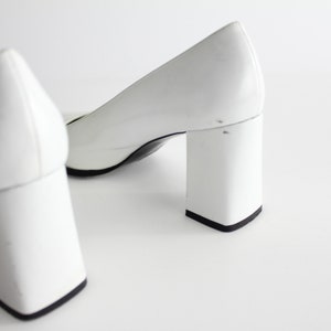 Puede incluir: Un par de zapatos de tac&oacute;n blanco con tac&oacute;n de bloque. Los zapatos est&aacute;n hechos de un material liso y brillante.
