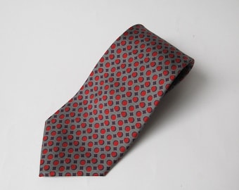 May include: A gray necktie with a repeating pattern of red circles and dark purple squares. The tie is angled across a white background, showcasing its design and texture. This accessory is suitable for formal or business attire.