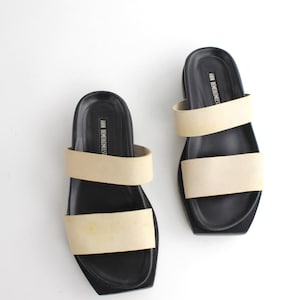 May include: A pair of black leather sandals with two beige straps. The sandals have a square toe and a thick platform sole. The brand name "ANN DEMEULEMEESTER" is printed on the straps.