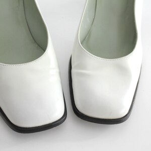 Puede incluir: Un par de zapatos de sal&oacute;n de cuero blanco con puntera cuadrada y ribete negro. Los zapatos est&aacute;n sobre un fondo blanco.