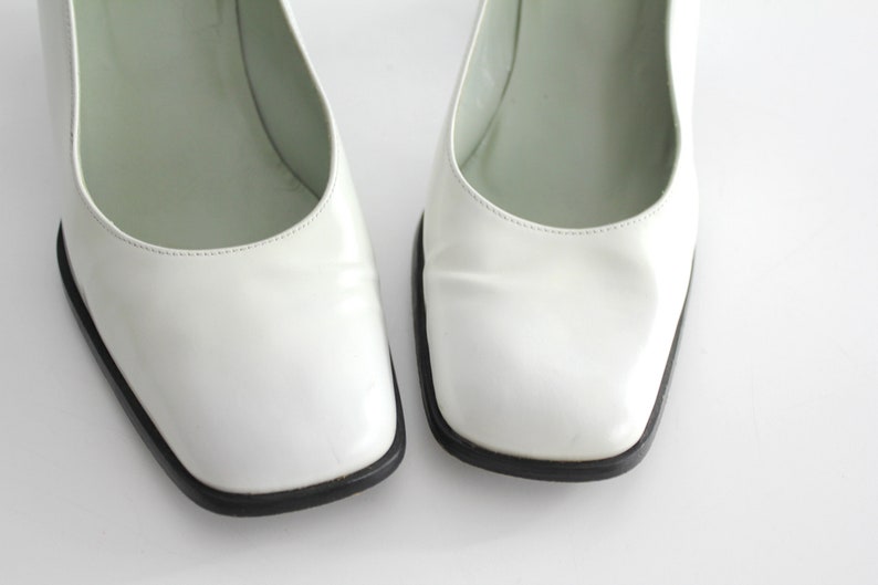 Puede incluir: Un par de zapatos de sal&oacute;n de cuero blanco con puntera cuadrada y ribete negro. Los zapatos est&aacute;n sobre un fondo blanco.
