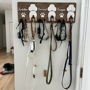May include: A wooden dog leash rack with three hooks, each labeled with a dog's name: Kodaline, Percival, and Reagan. Six leashes hang from the hooks, including a white leash with a small pouch.