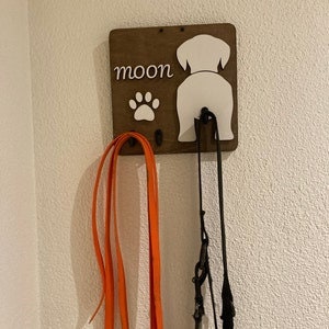 May include: Wooden dog leash holder with a white silhouette of a dog's rear end and the word "moon" written above it. A paw print is below the word "moon". Two leashes are hanging from the holder.