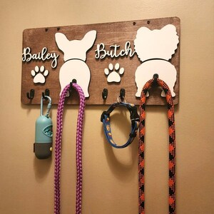 May include: A wooden dog leash and collar rack with two dog silhouettes, the names "Bailey" and "Butch" are written on the rack, and there are paw prints below each name. There are three leashes and a collar hanging on the rack.