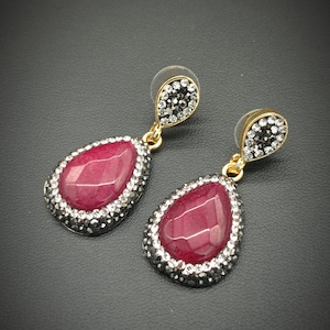 Può includere: Un paio di orecchini a goccia. Ogni orecchino presenta una grande pietra rosso rubino sfaccettata, circondata da un bordo di piccoli cristalli trasparenti. La parte superiore ha un gruppo di cristalli neri e trasparenti, con accenti metallici dorati.