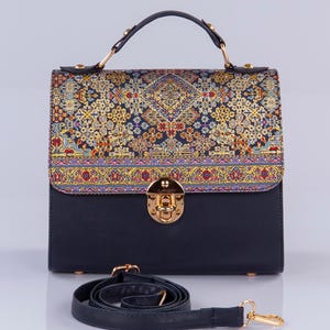 Op de afbeelding: Een zwarte handtas met een gouden sluiting en een gepatenteerde klep. De klep heeft een kleurrijk ontwerp dat doet denken aan een Perzisch tapijt. De tas wordt geleverd met een verstelbare zwarte schouderband.