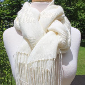 Puede incluir: Bufanda tejida de color crema con un patrón texturizado y flecos. La bufanda está drapeada sobre un maniquí blanco, mostrando su diseño y textura. Los flecos cuelgan en la parte inferior de la bufanda.