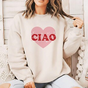 May include: A cream-colored sweatshirt featuring a pink heart with the word "CIAO" in red lettering. The heart design includes small snowflake accents. The sweatshirt has long sleeves and a crew neck.