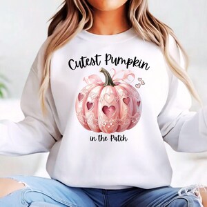 May include: White sweatshirt featuring a pink pumpkin design with heart-shaped cutouts and the text "Cutest Pumpkin in the Patch." The pumpkin has a bow and is decorated with hearts. A fall-themed graphic.