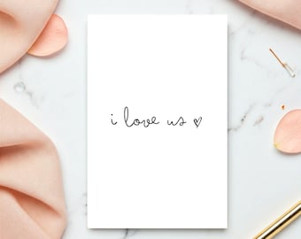 May include: A white card with the handwritten text "i love us" and a heart symbol. The card is surrounded by a pink fabric, a gold pen, and a marble surface.