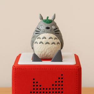 May include: A red cube-shaped speaker with a Totoro figure on top. The speaker has a textured surface and a pattern of small holes forming a silhouette. The Totoro figure is gray with a white belly and a green leaf.