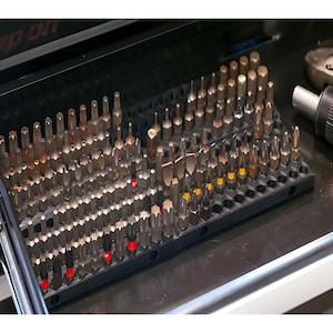 May include: A black plastic tray filled with a variety of screwdriver bits. The bits are organized in rows and columns, and they are all different sizes and shapes. The tray is sitting on a black surface.