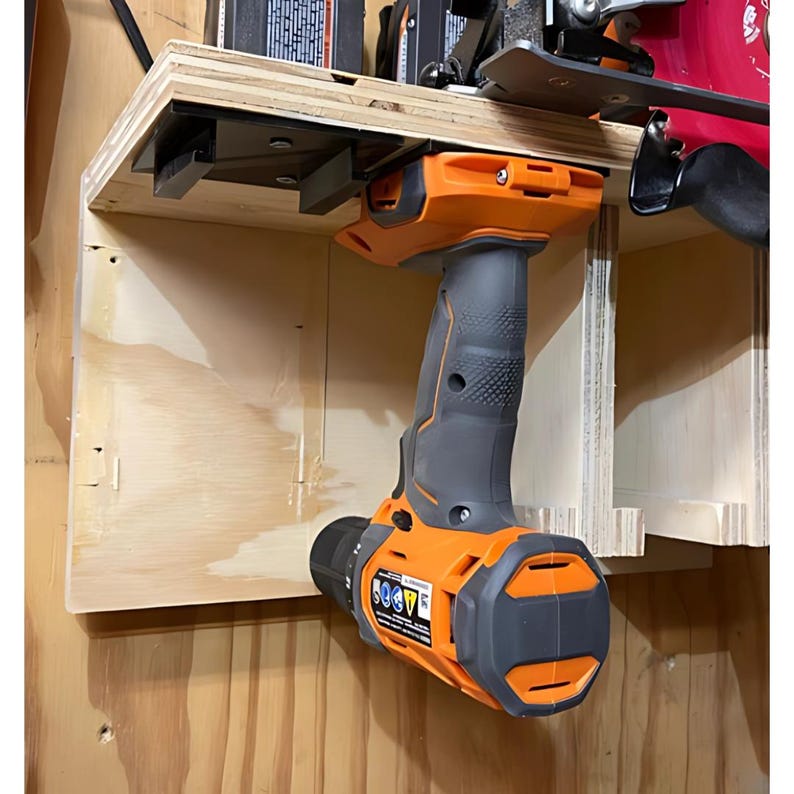 May include: A black and orange cordless drill is mounted on a wooden wall shelf. The drill is hanging upside down with the handle facing up. The drill is attached to the shelf with a black metal bracket.