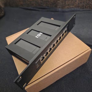 Könnte beinhalten: Ein schwarzer NETGEAR Netzwerk-Switch mit mehreren Anschlüssen, der auf einem braunen Karton liegt. Der Switch trägt den Text "TGE" auf der Oberseite und "NETGEAR" auf der Frontplatte. Er ist für die Netzwerkverbindung konzipiert.