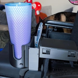 May include: A black wheelchair accessory with a cup holder and a phone holder. A blue and purple textured tumbler is in the cup holder. A clear phone is in the phone holder. The accessory is attached to a black wheelchair.