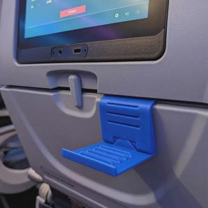 May include: A blue plastic phone holder attached to the back of an airplane seat. The holder is folded down, ready to hold a phone or tablet. The screen in front displays text that says "PA in progress. Please remove your headphones."