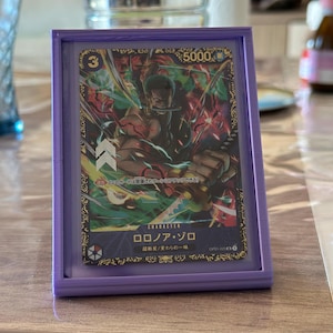 May include: A framed trading card featuring a character illustration with Japanese text. The card is set in a purple frame. The card's design includes a character illustration, the number 3, and the number 5000. The frame is on a wooden surface.