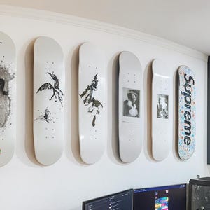 May include: Six skateboards displayed on a white wall. The boards showcase diverse artwork, including abstract designs, a portrait, and the word "Supreme" in a floral pattern. The skateboards are arranged in a line.