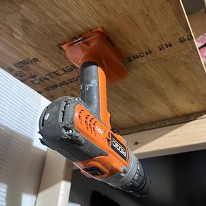 May include: An orange and black cordless drill with the brand name "RIDGID" and the text "18V" and "20" printed on the side. The drill is hanging upside down from a wooden surface.