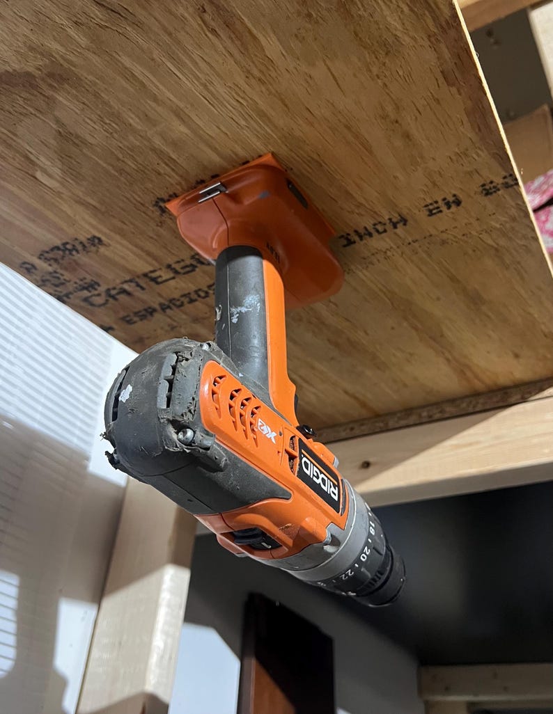 May include: An orange and black cordless drill with the brand name "RIDGID" and the text "18V" and "20" printed on the side. The drill is hanging upside down from a wooden surface.