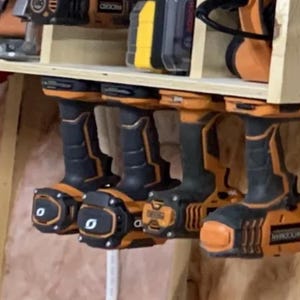 May include: Four orange and black cordless drills hanging on a wooden wall rack. The drills are all different brands and have different features.
