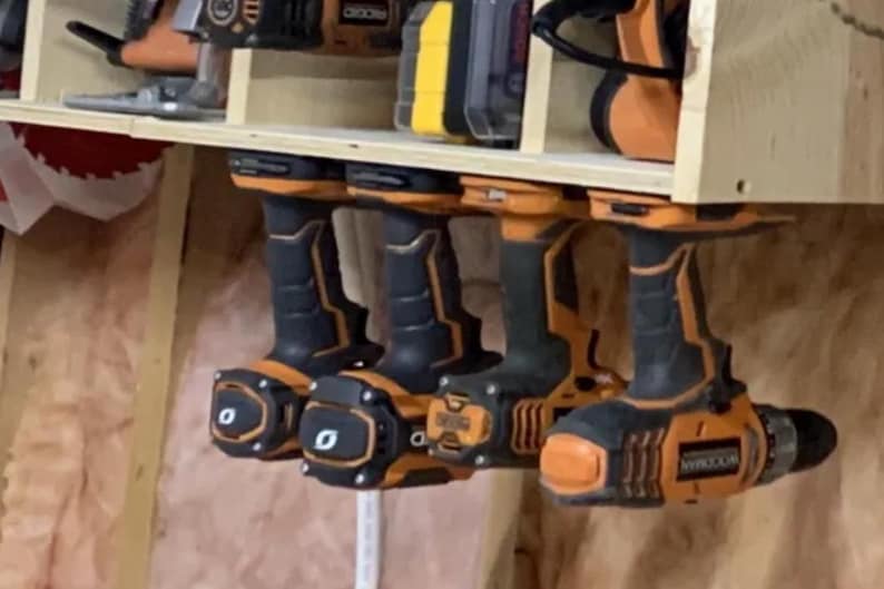 May include: Four orange and black cordless drills hanging on a wooden wall rack. The drills are all different brands and have different features.