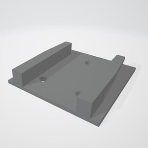 May include: A gray plastic wall mount with two holes for mounting screws. The mount has a raised lip on one side to hold a device in place.