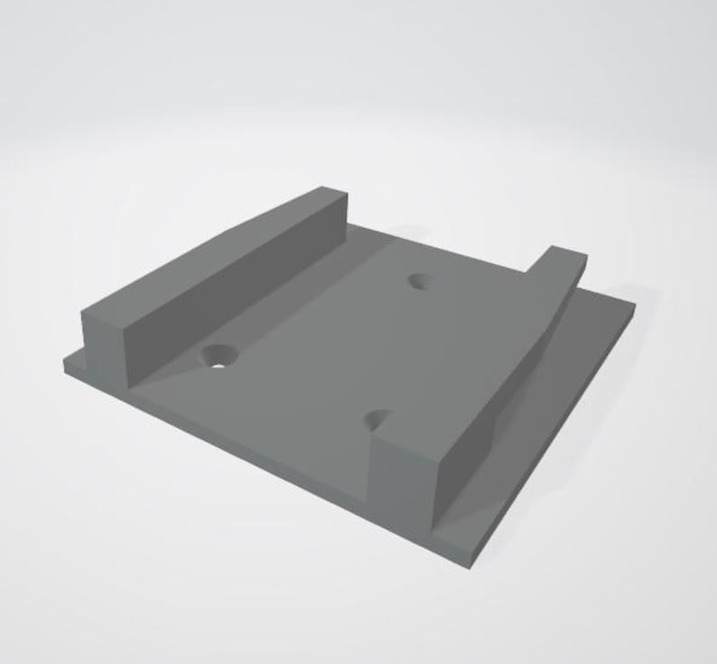 May include: A gray plastic wall mount with two holes for mounting screws. The mount has a raised lip on one side to hold a device in place.