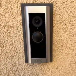 May include: A silver Ring doorbell mounted on a beige stucco wall. The doorbell has a black camera lens and a black button.
