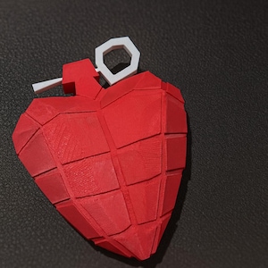 Peut inclure: Objet rouge en forme de cœur avec un design géométrique, ressemblant à une grenade. Il a une goupille blanche et une goupille rouge. L'objet est posé sur un fond sombre.