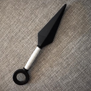 May include: A black kunai knife with a white wrapped handle and a circular ring at the base. The blade is a dark, matte black, and the handle is wrapped in white material. The object is set against a textured grey background.