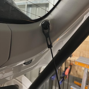 May include: Close-up of a car's interior, focusing on the rear window and its supporting components. A black plastic latch is attached to the white interior panel, with a black cord extending downwards. The rear window has a black border and thin defroster lines.