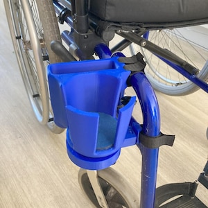 May include: A blue cup holder attached to a wheelchair. The cup holder is made of plastic and has a cylindrical base and a rectangular cup holder. The wheelchair has a black seat and blue frame.