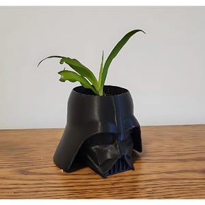 May include: A black Darth Vader planter with a green plant growing inside. The planter is made of plastic and has a detailed design.