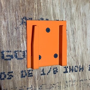May include: Orange plastic wall mount with two slots for holding objects. The mount is attached to a wood surface with screws.