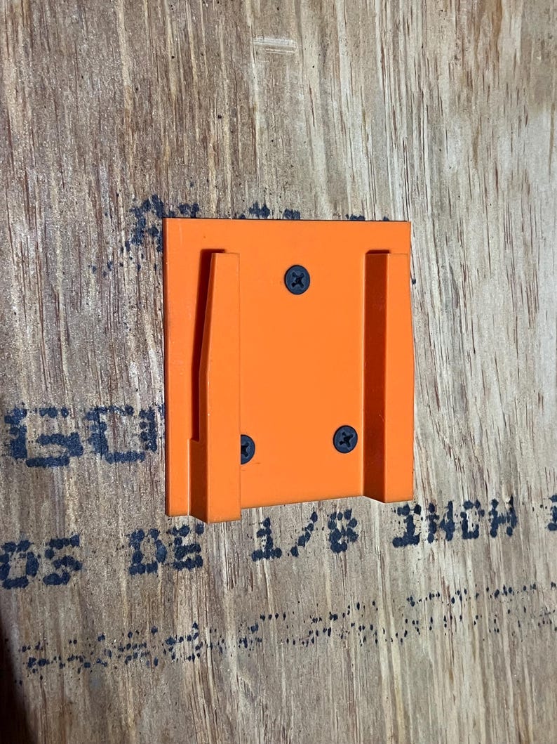 May include: Orange plastic wall mount with two slots for holding objects. The mount is attached to a wood surface with screws.