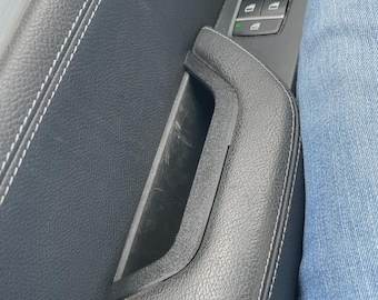 May include: Close-up of a black car door panel with white stitching. The door handle is integrated into the panel. Window control buttons are visible. A person's blue jeans are in the frame.