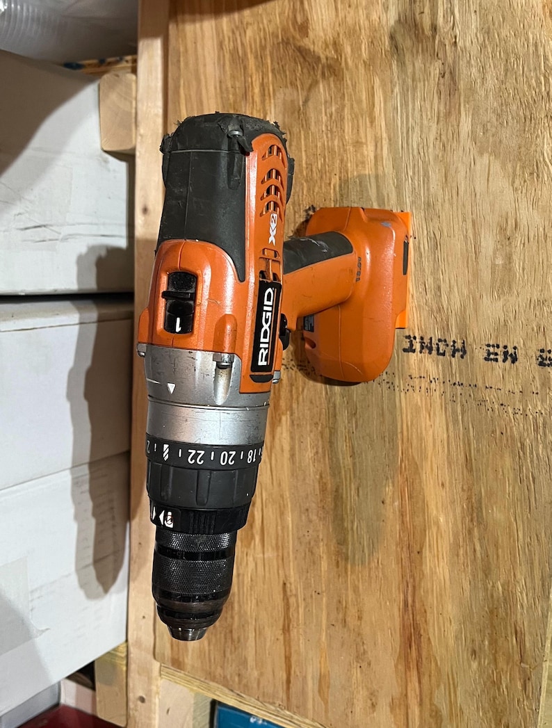 May include: An orange and black cordless drill with a black chuck and a silver dial with numbers 18 to 22. The drill is mounted on a wooden surface.