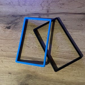 May include: Two rectangular frames, one bright blue and one black, are on a wooden surface. The blue frame stands upright, while the black frame leans against it, creating a geometric composition. The frames appear to be made of plastic.