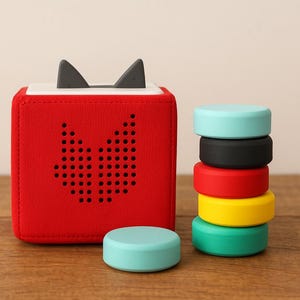 May include: A red fabric cube with black speaker holes and grey cat ears. A stack of colourful, round blocks in teal, black, red, yellow, and green sits beside the cube. A teal block rests on the wooden surface.