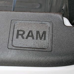 May include: Close-up of a textured black surface with the embossed word "RAM" in a rectangular frame. The image shows a portion of a vehicle's exterior, with a white edge visible at the bottom.