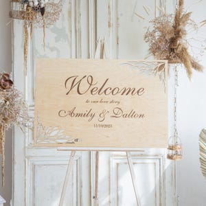 Könnte beinhalten: Ein hölzernes Willkommensschild auf einer Staffelei. Das Schild trägt das Wort "Welcome" in eleganter Schrift, gefolgt von "to our love story" und den Namen "Amily & Dalton" mit dem Datum "11/10/2023". Dekorative ausgeschnittene Blumendesigns schmücken die Ecken.