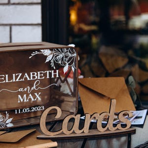 Caja personalizada para tarjetas de boda con diseño floral y cartel de madera para una recepción rústica.