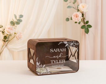 Pode incluir: Uma caixa de madeira retangular para cartões com um painel frontal de acrílico transparente com arte floral branca e o texto "SARAH and TYLER 02/06/2026". Os lados da caixa castanho-escura têm recortes em forma de envelope e uma ranhura.