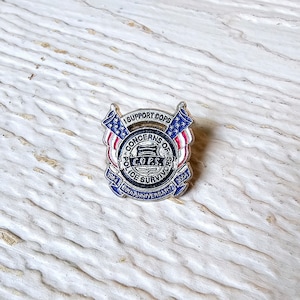 May include: A silver-toned pin with a patriotic design. The pin features the text "I SUPPORT COPS" above a central emblem with "CONCERNS OF POLICE SURVIVORS" and "C.O.P.S." The pin commemorates the 20th anniversary, 1984-2004.